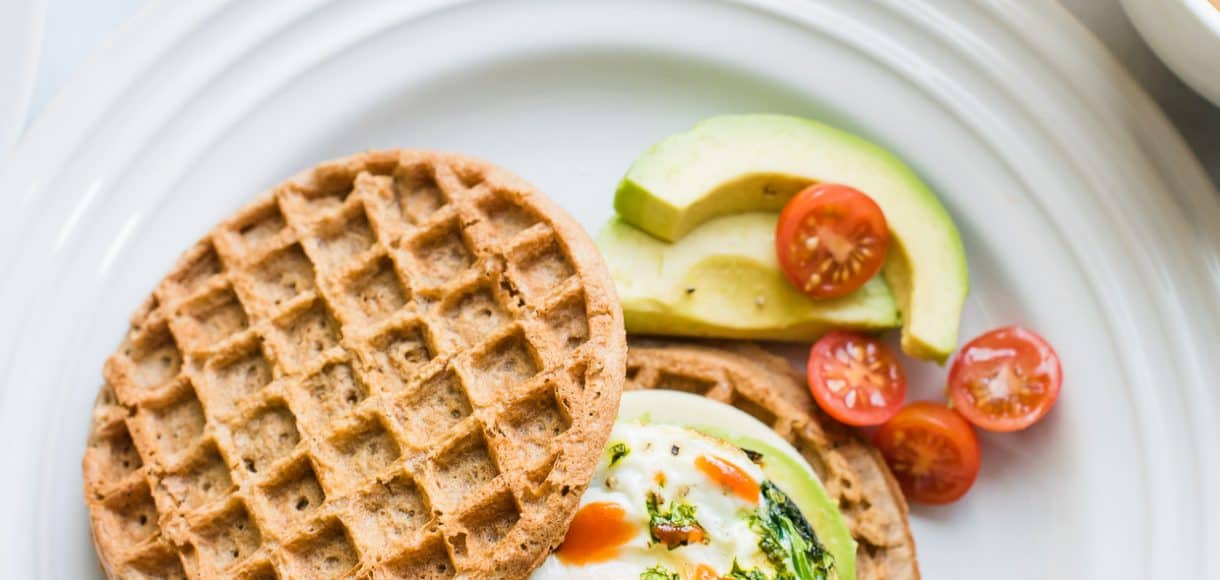 Two wholegrain waffles, one topped with an over easy egg, spinach, avocado, and hot sauce to form a Waffle Breakfast Sandwich.