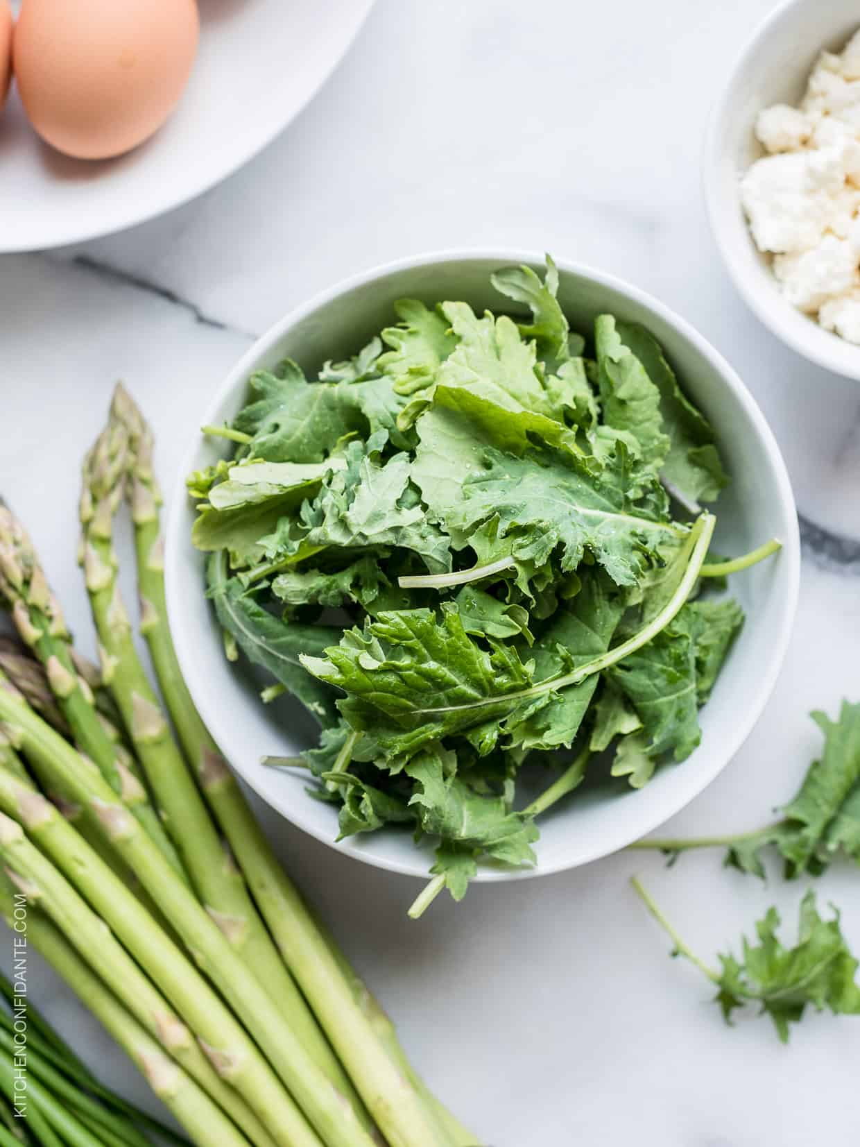 A white bowl filled with fresh kale alongside stalks of asparagus. 