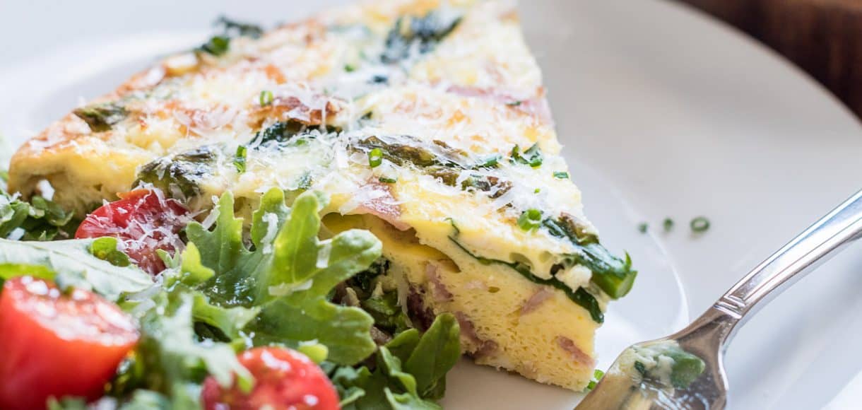 A wedge of Asparagus, Ham and Kale Frittata served on a white plate alongside a green salad.