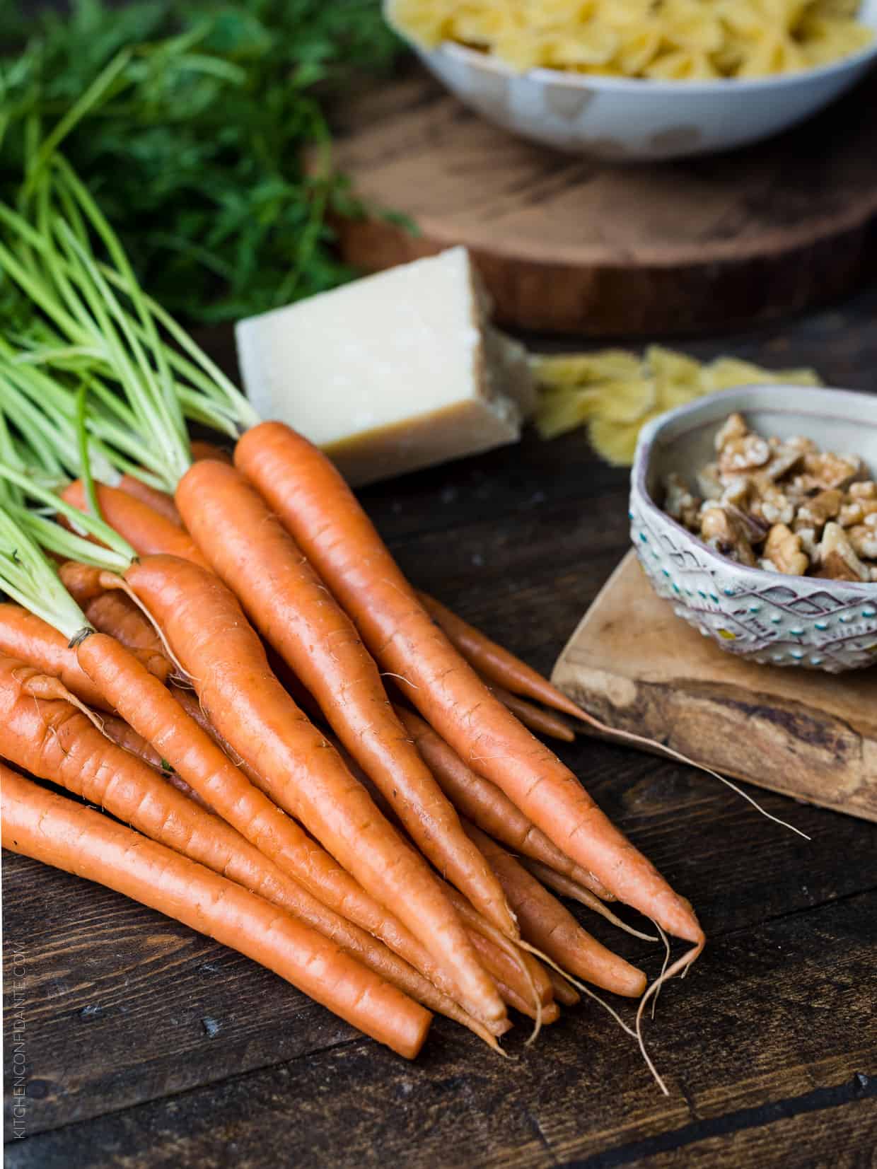 A bunch of raw carrots on a wooden surface surrounded by a block of Parmesan and a bowl of walnuts.