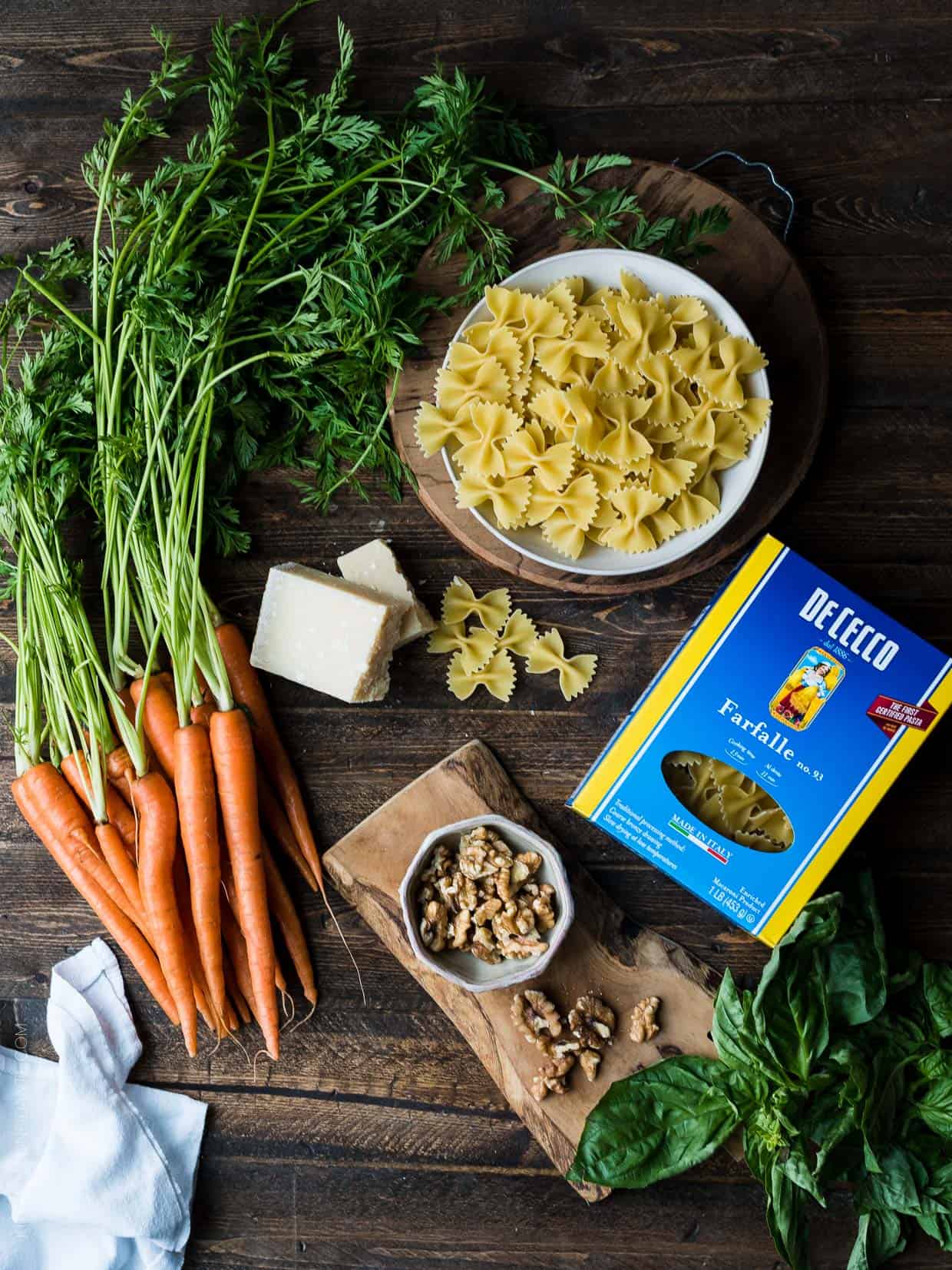 A wooden surface topped with all the ingredients needed to make Farfalle with Roasted Carrot Pesto, Parmesan and Toasted Walnuts.