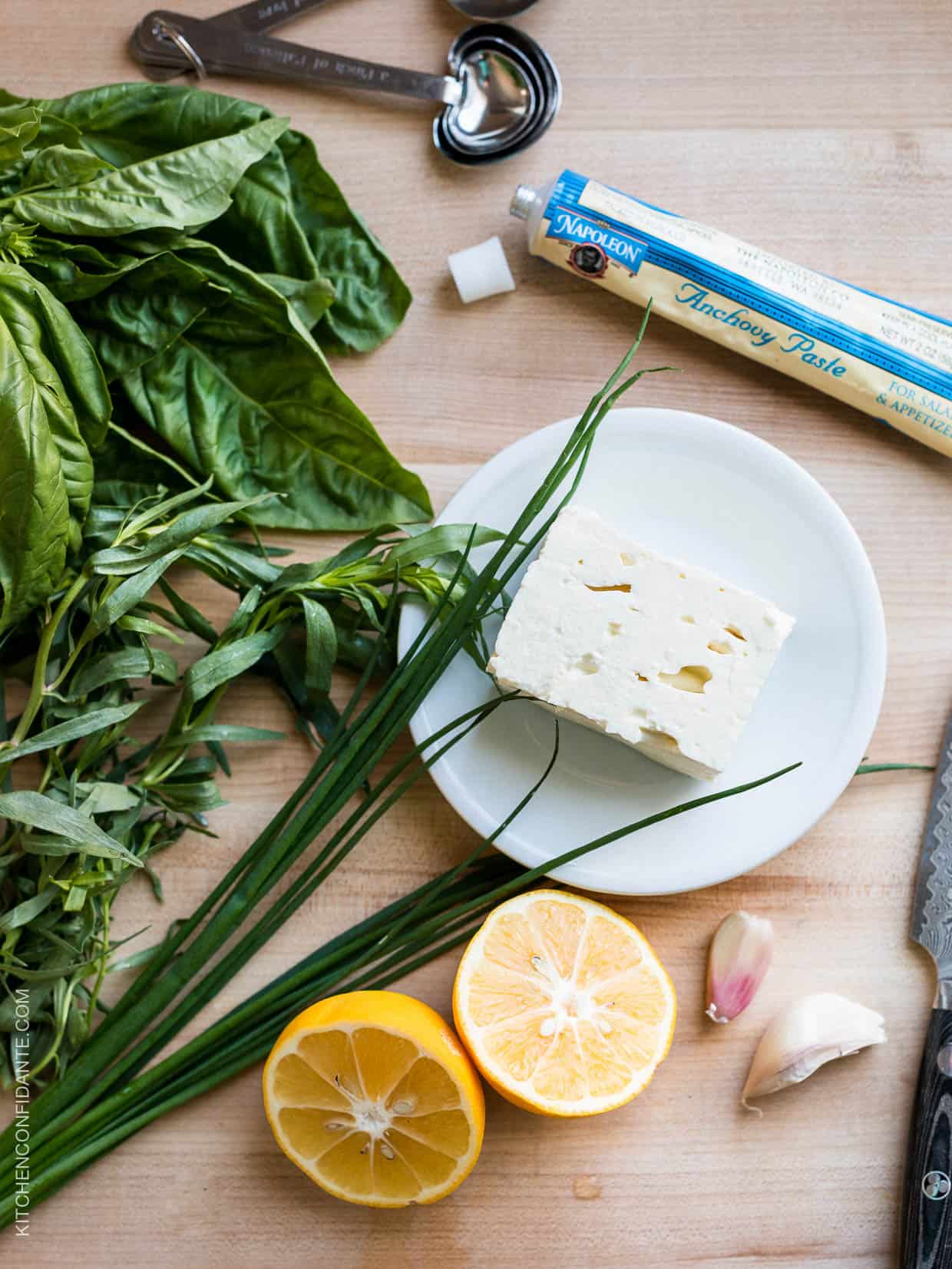 Fresh herbs, a halved lemon, and a small block of feta cheese on a wooden surface.