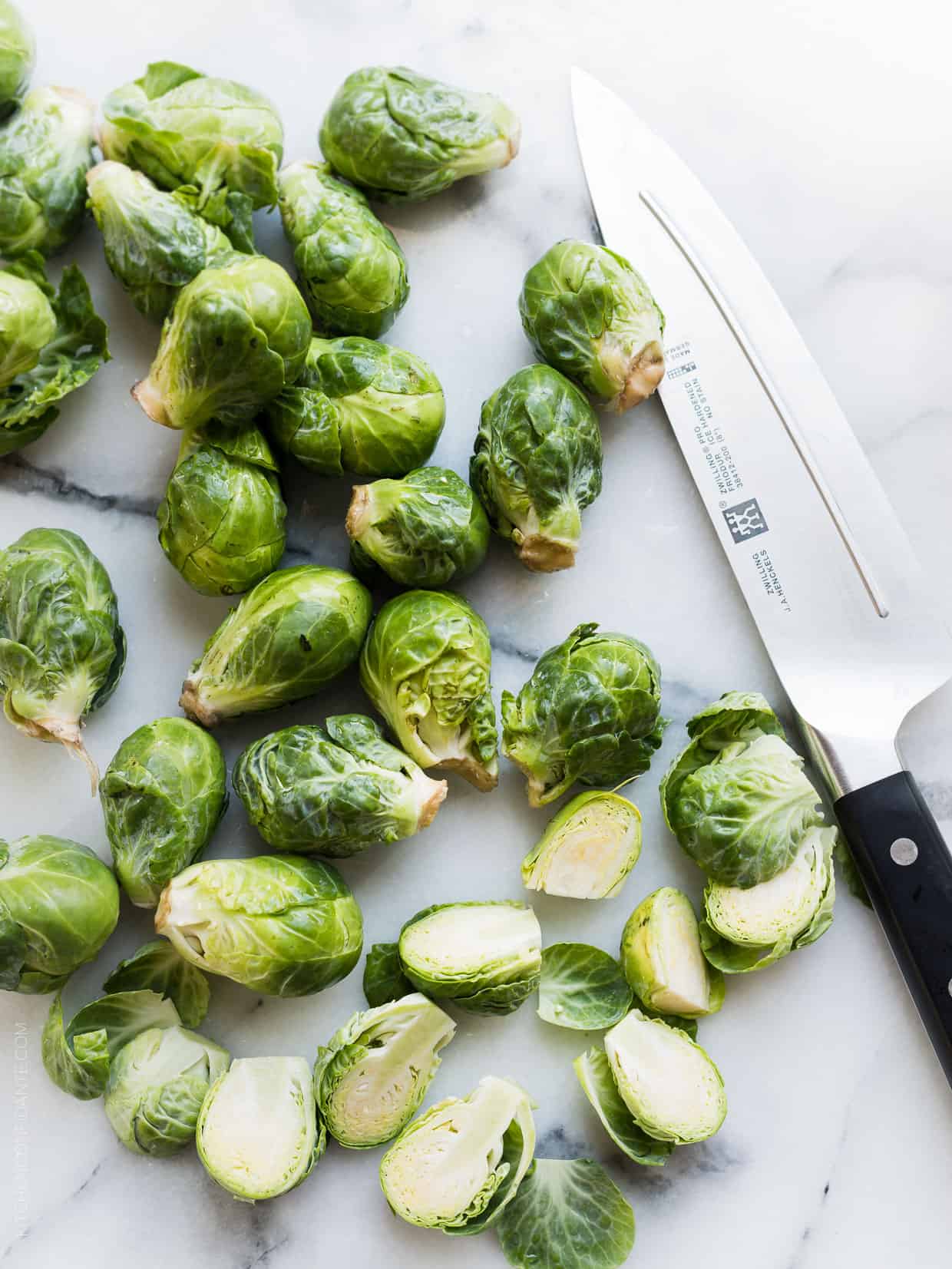 Brussels sprouts split in half on a marble surface alongside a chef's knife.