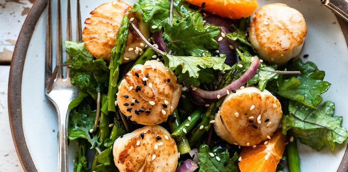 Pan Seared Scallops in a white dish over a bed of kale, asparagus, and fresh slices of tangerine.