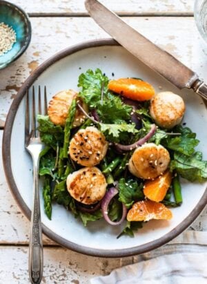 Pan Seared Scallops in a white dish over a bed of kale, asparagus, and fresh slices of tangerine.