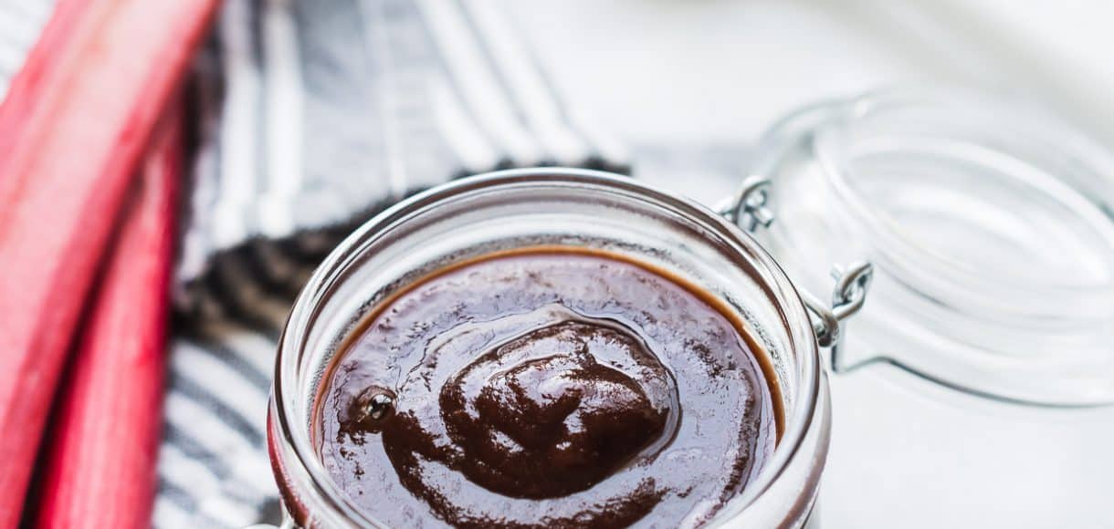 Glass jar of Rhubarb-Wine BBQ sauce surrounded by fresh stalks of rhubarb.