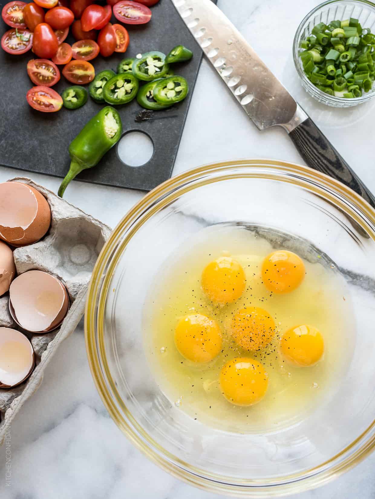 Six eggs cracked into a glass bowl surrounded by chopped peppers and tomatoes.