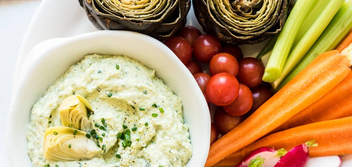 A bowl of Artichoke and Avocado Dip surrounded by vegetables including carrots, radishes, and fresh artichokes.