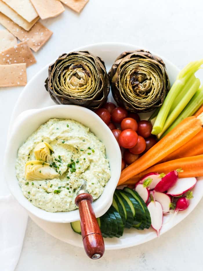 A bowl of Artichoke and Avocado Dip surrounded by vegetables including carrots, radishes, and fresh artichokes.