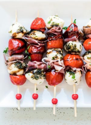 A row of Peppadew Pepper Caprese Skewers with Balsamic Glaze made with fresh mozzarella and prosciutto on a white plate.