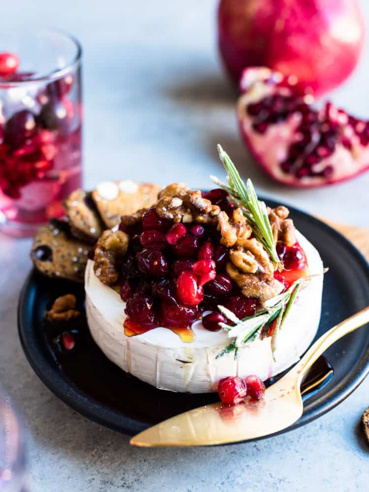 Cranberry Pomegranate Baked Brie served on a black plate with segments of fresh pomegranate in the background.