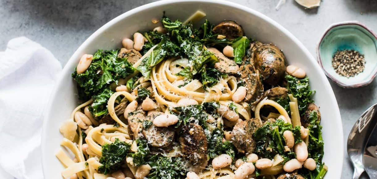 Fettuccine with Chicken Sausage, Kale and Cannellini Beans piled high in a white bowl with serving utensils alongside.