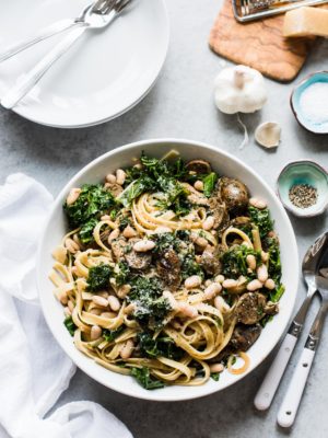 Fettuccine with Chicken Sausage, Kale and Cannellini Beans piled high in a white bowl with serving utensils alongside.