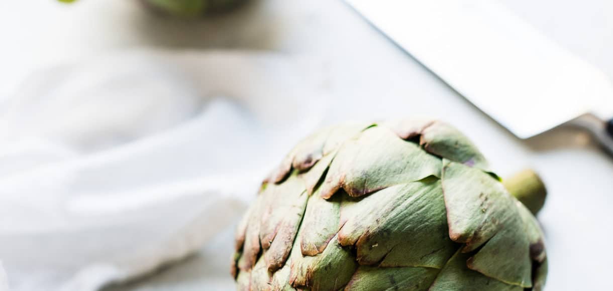 Two artichokes and a knife on a cutting board.