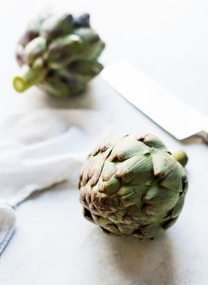 Two artichokes and a knife on a cutting board.
