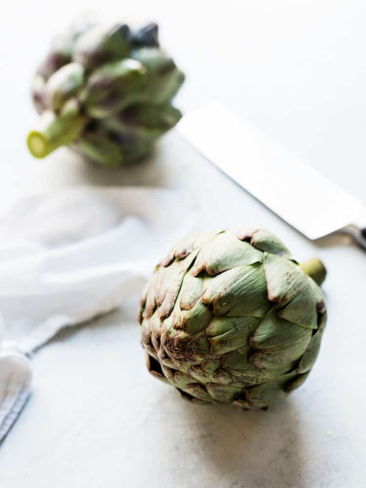 Two artichokes and a knife on a cutting board.