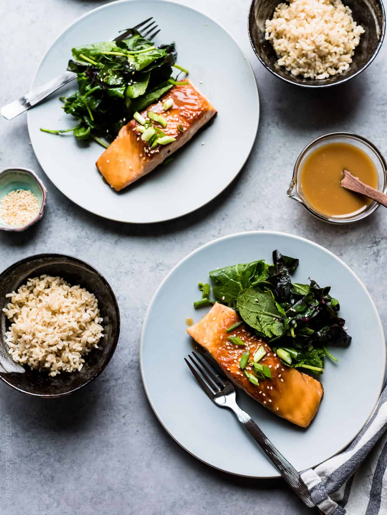 Two white plates filled with Miso Maple Glazed Salmon and wilted green salads served alongside bowls of rice.