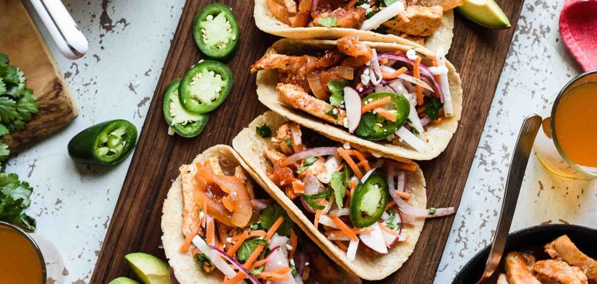 Chipotle Chicken Tacos with Jicama Slaw on a wooden board with avocados, jalapeños and radishes.