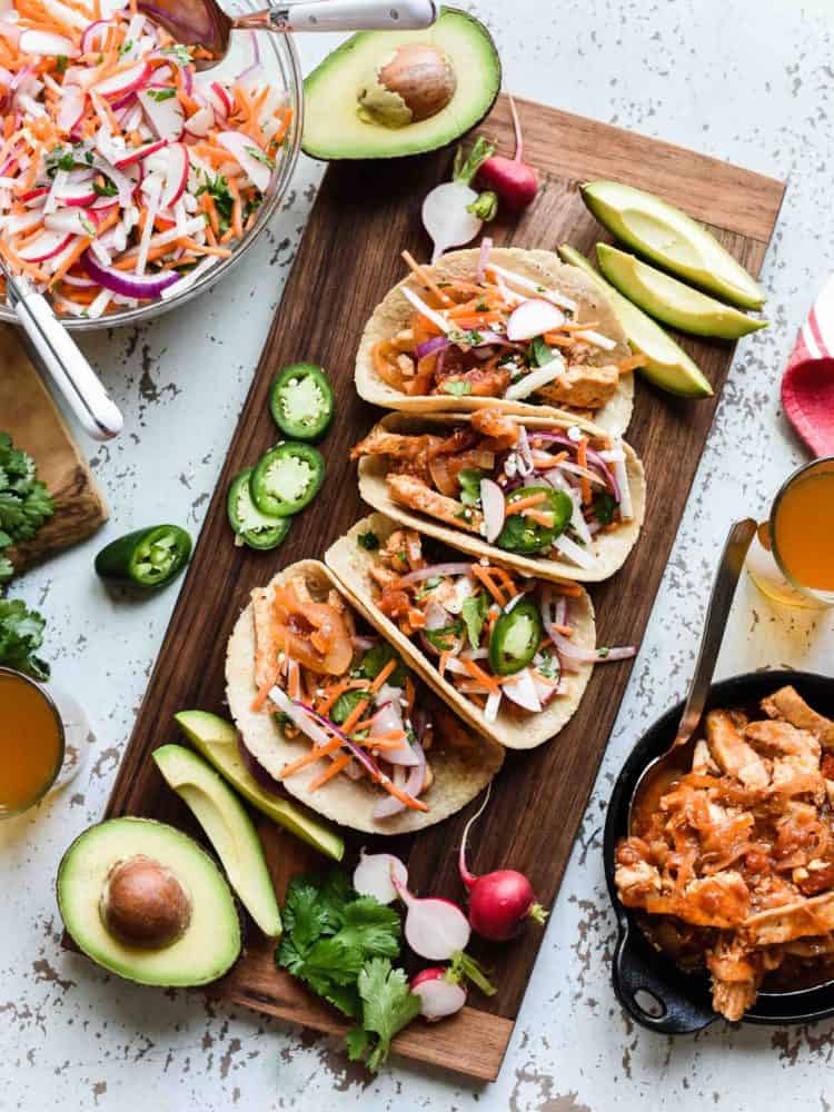 Chipotle Chicken Tacos with Jicama Slaw on a wooden board with avocados, jalapeños and radishes.