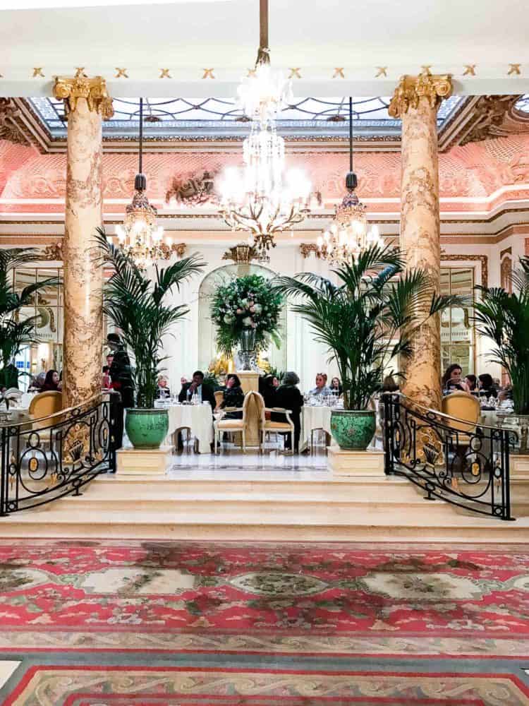 An interior shot of the Ritz in London with pink Oriental rugs, pink marble, ornate chandeliers, and large green potted plants.