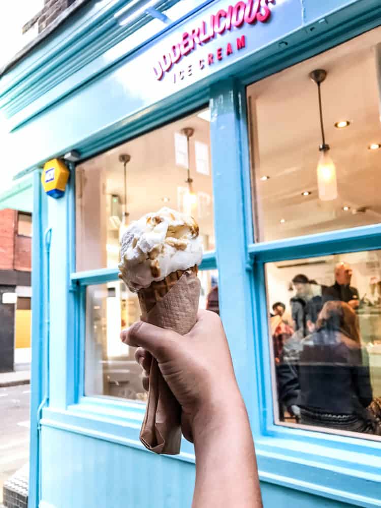 A hand holding an ice cream cone in front of a turquoise ice cream store front.