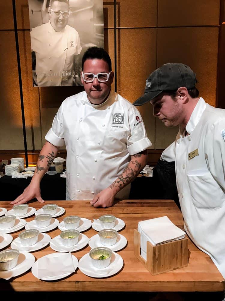 Two men in white chefs coats standing by small plates of food on a wooden table at the Pebble Beach Food & Wine.