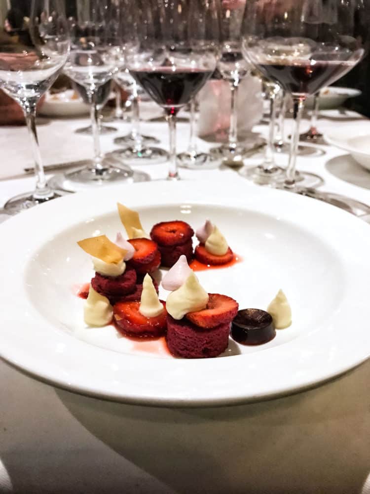Strawberries, red velvet cake, and frosting on a white plate with wine glasses in the background at the Pebble Beach Food & Wine.