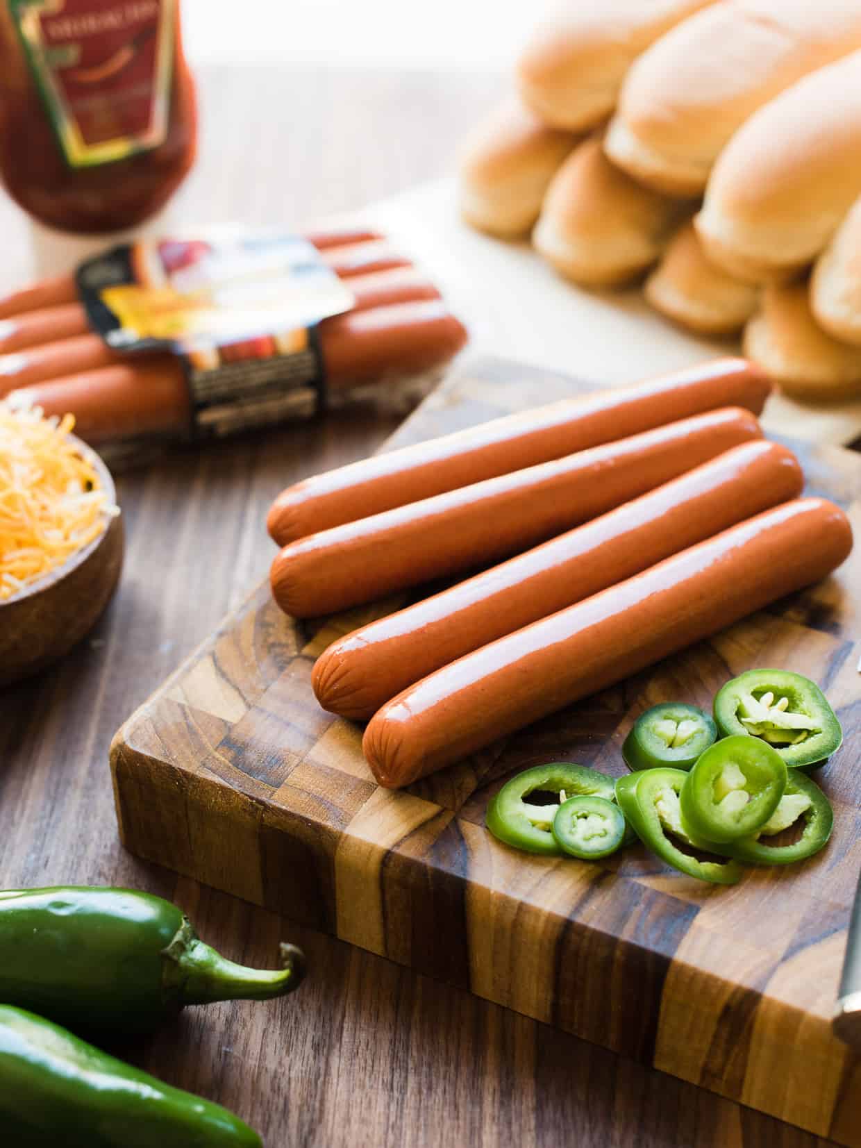 Hot dogs, buns, cheese, jalapeños, and Sriracha ketchup on a wooden board.