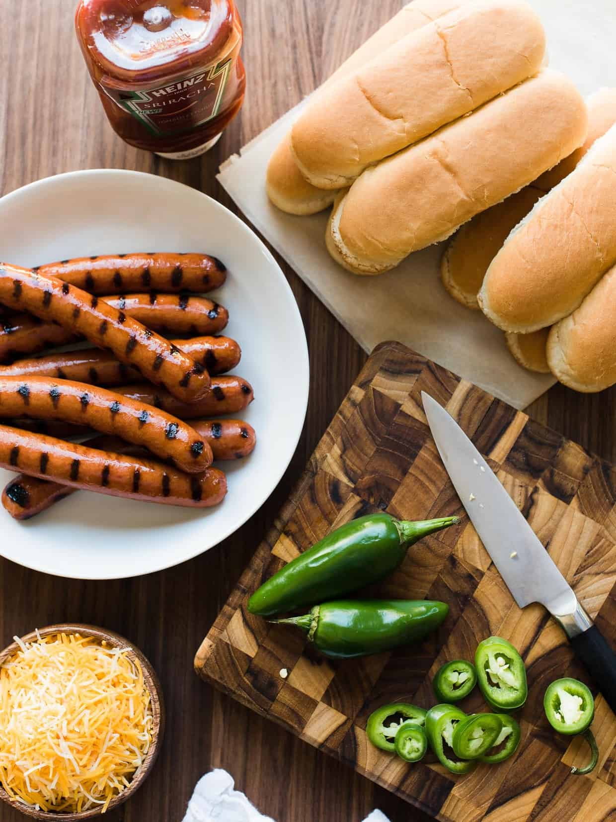 Grilled hot dogs, buns, cheese, jalapeños, and Sriracha ketchup on a wooden board.