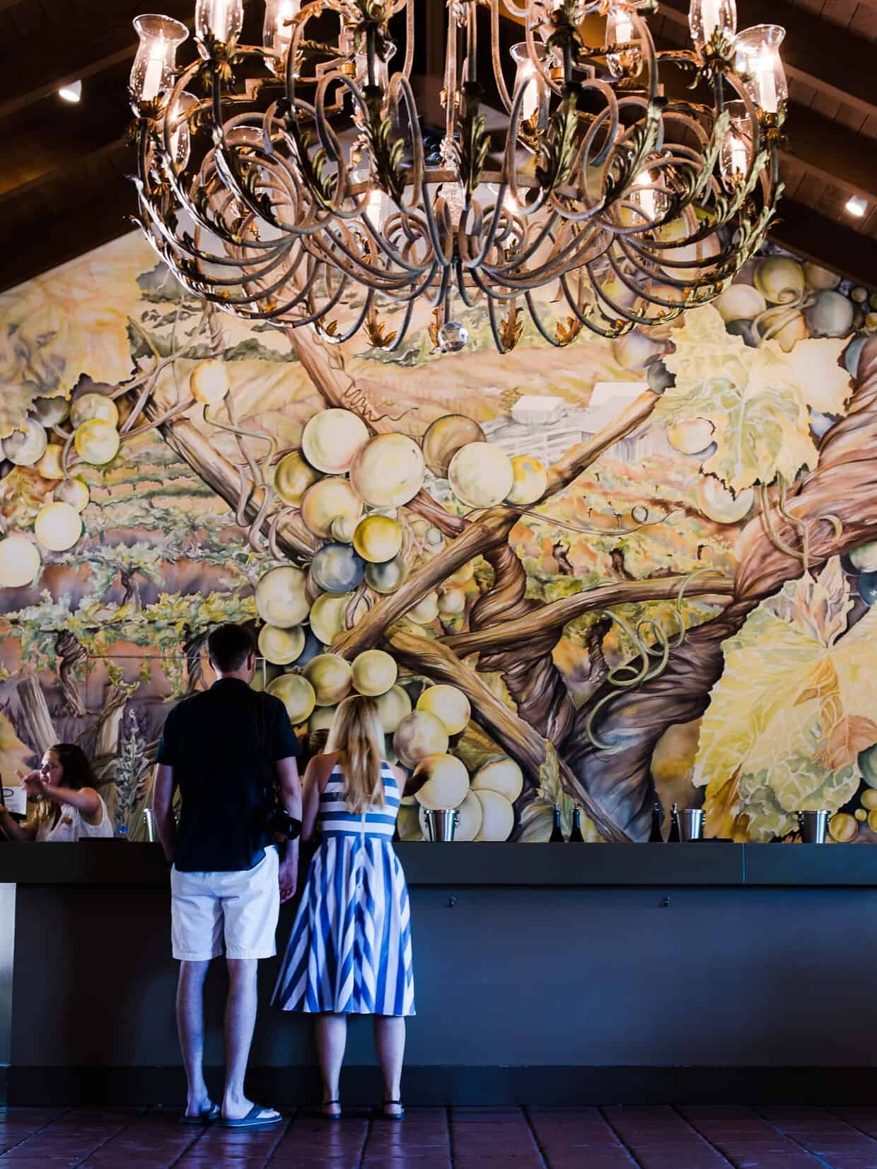 A man and a woman standing in front of a large mural under a large chandelier. 