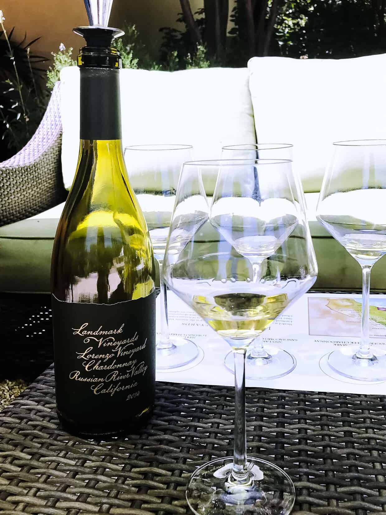 A bottle of wine next to wine glasses on a wicker table outside.