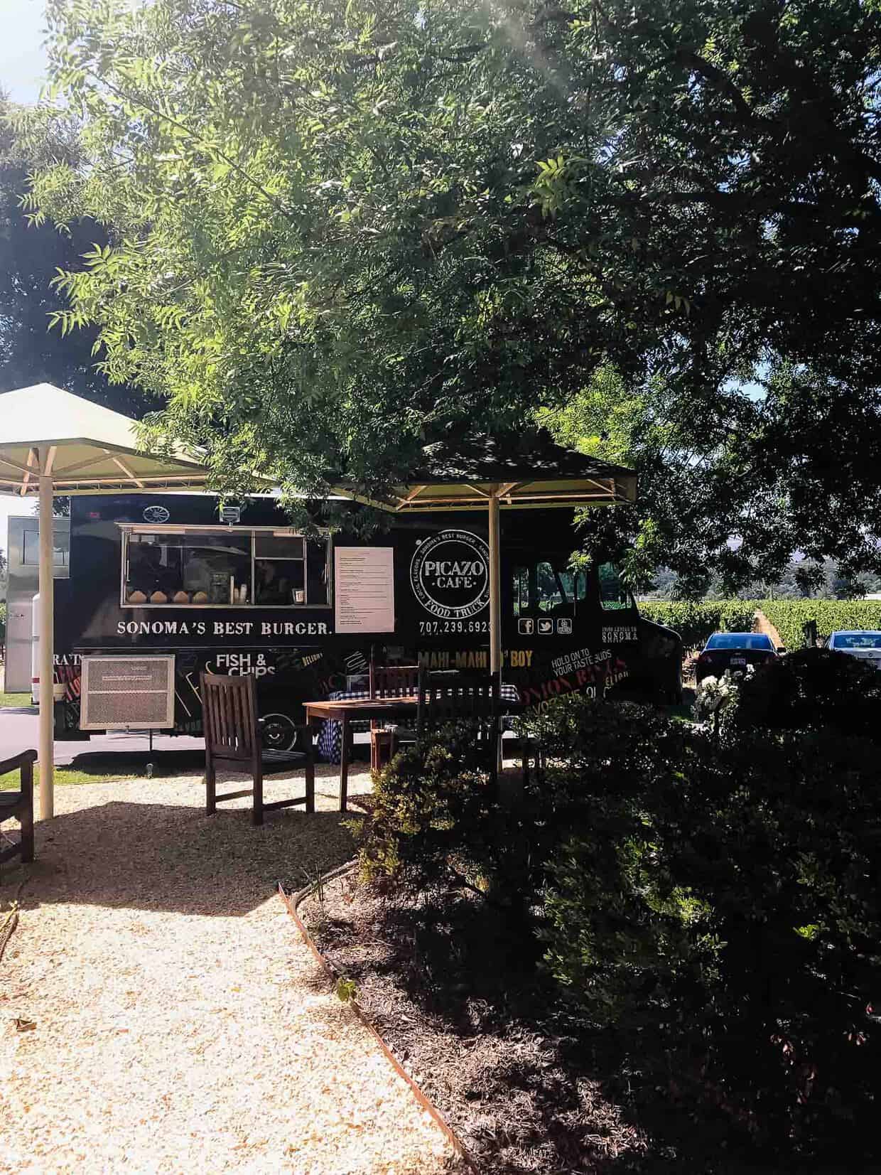 A black and white food truck parked under the shade of trees with outdoor seating.