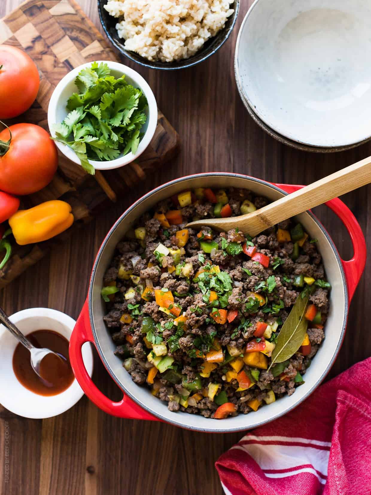 Red dutch oven of Filipino-style Picadillo surrounded by fresh ingredients and brown rice.