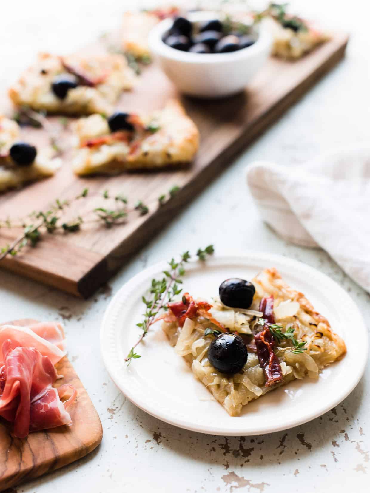 Slice of homemade Pissaladière with Prosciutto on white plate with more slices on a wooden serving board in the background.