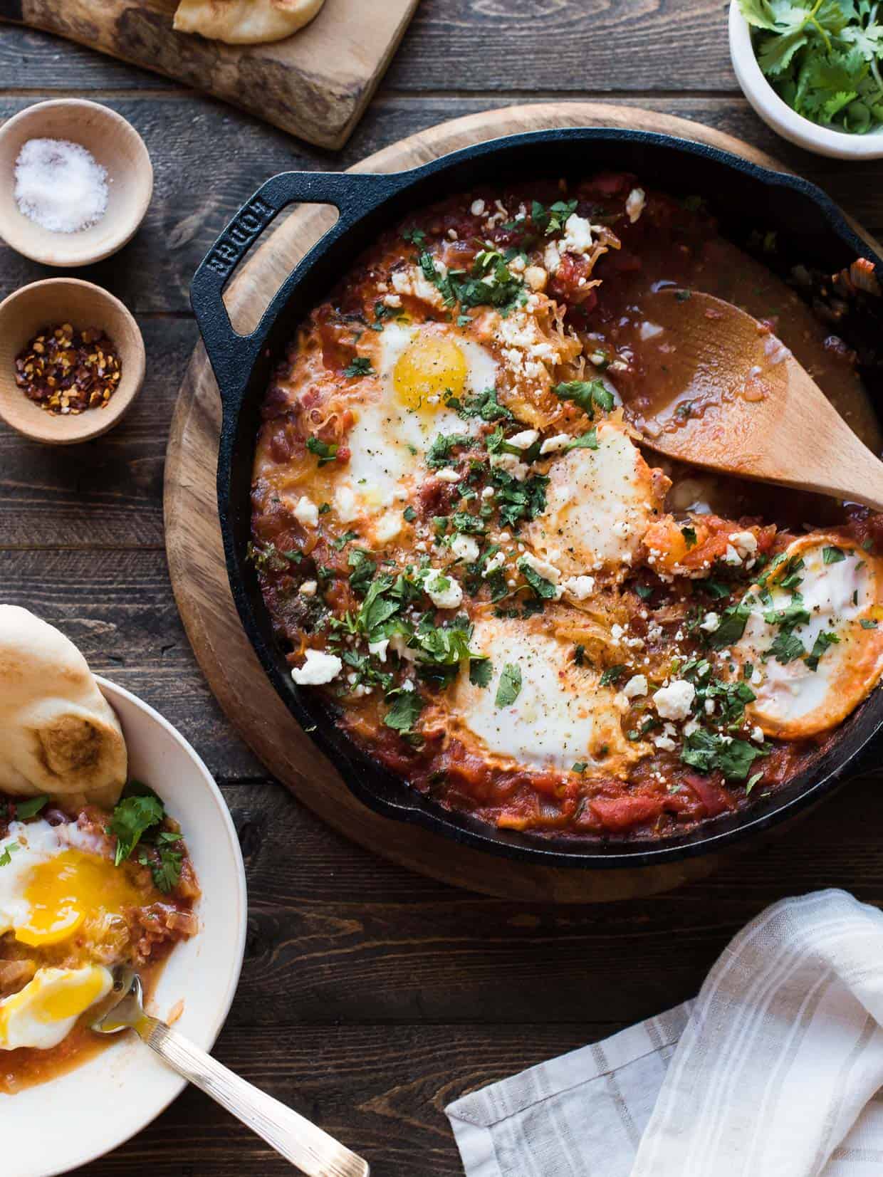 Cast iron pan filled with Spaghetti Squash Shakshuka and surrounded with garnishes and seasonings.