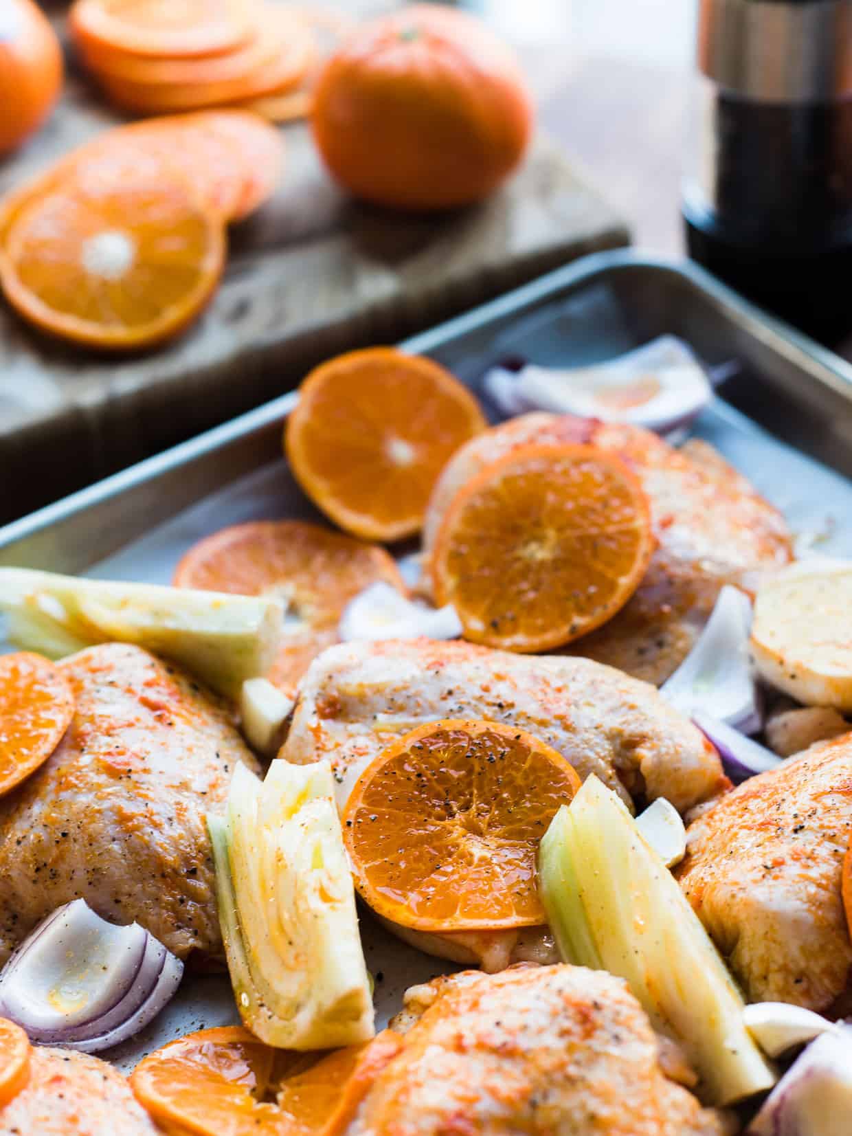 Preparing chicken thighs with mandarin orange slices, fennel, red onion on baking sheet.