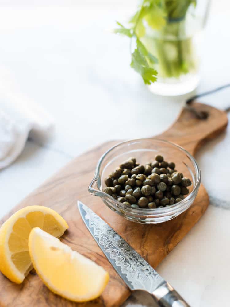 Glass dish with capers on a cutting board with lemon slices.