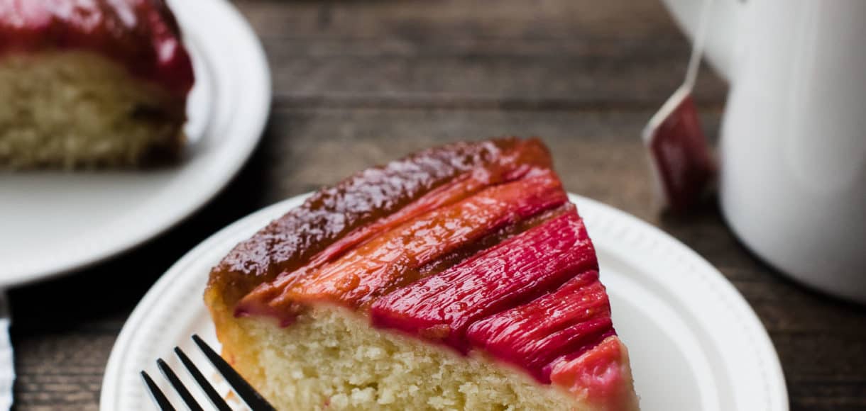 Slice of Rhubarb Upside Down Cake on a dessert plate with a fork and a mug of tea in background.