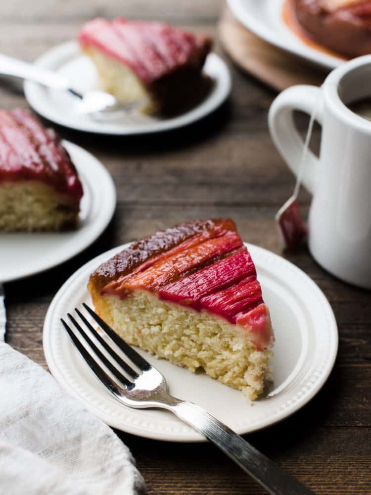 Slice of Rhubarb Upside Down Cake on a dessert plate with a fork and a mug of tea in background.