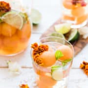 A glass of white sangria with cantaloupe, lime, mint and edible flowers.