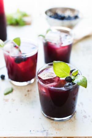 Glasses of blueberry ginger basil soda garnished with basil and blueberries.