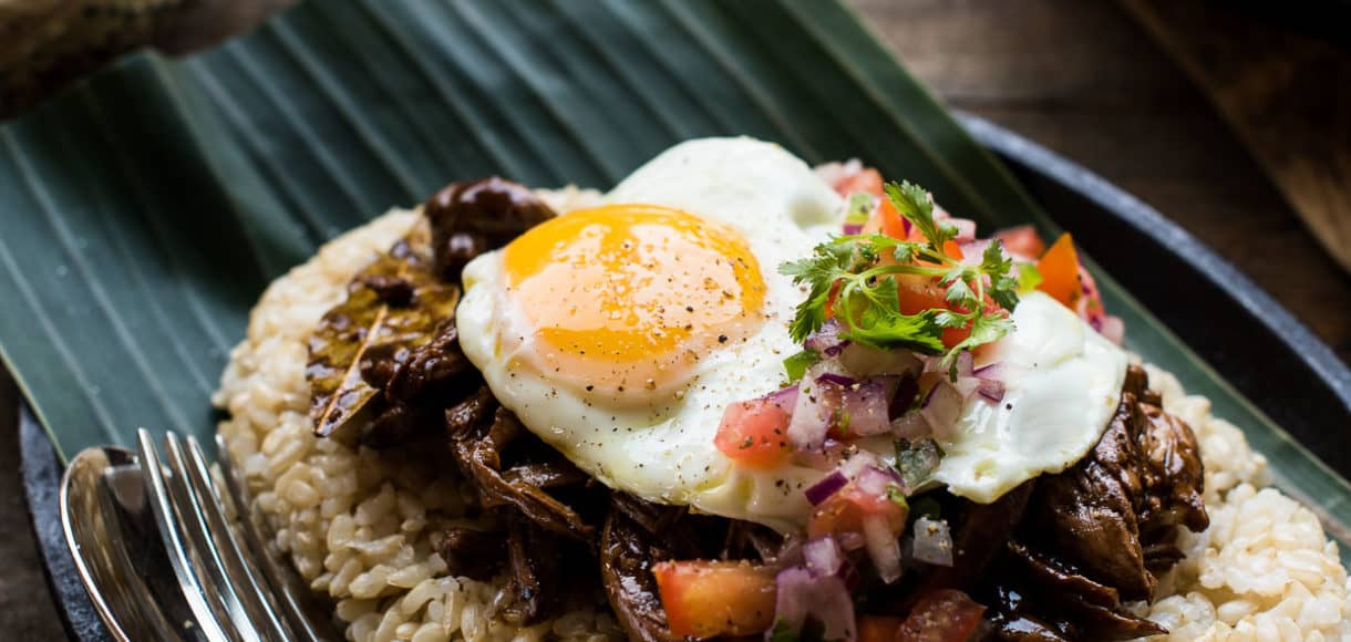 Adobo Loco Moco with shredded chicken adobo on a bed of brown rice with a sunnyside up egg, tomatoes, onions, on a banana leaf.
