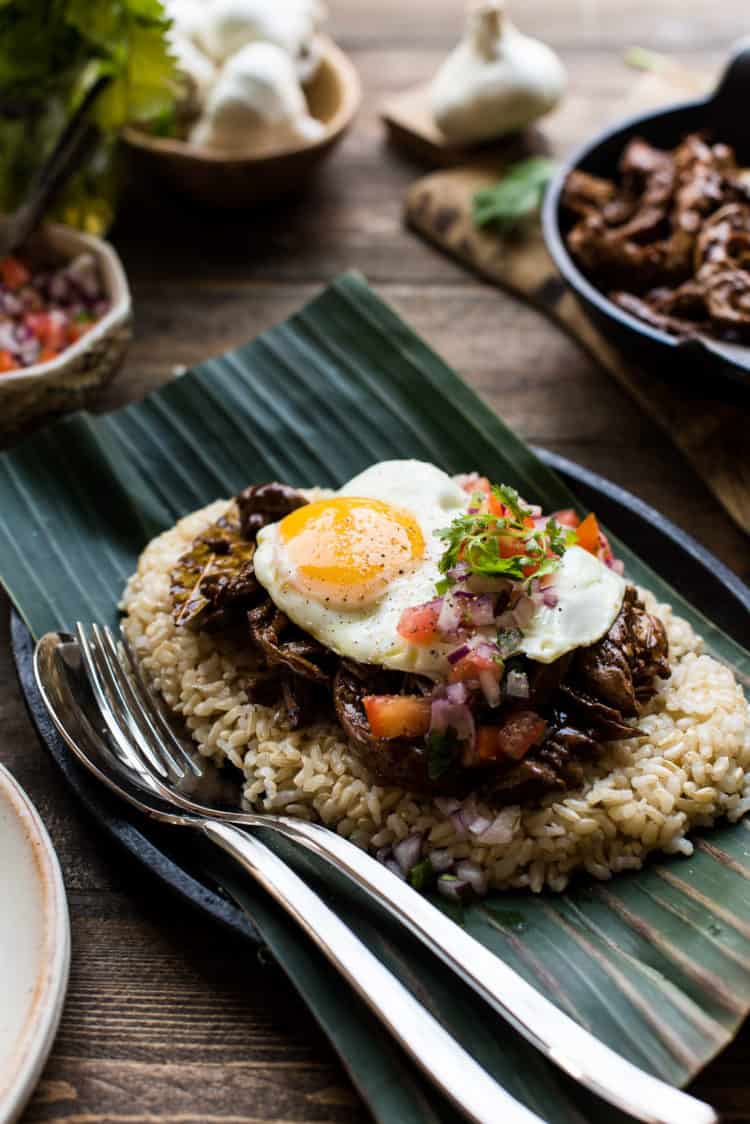 Adobo Loco Moco with shredded chicken adobo on a bed of brown rice with a sunnyside up egg, tomatoes, onions, on a banana leaf.