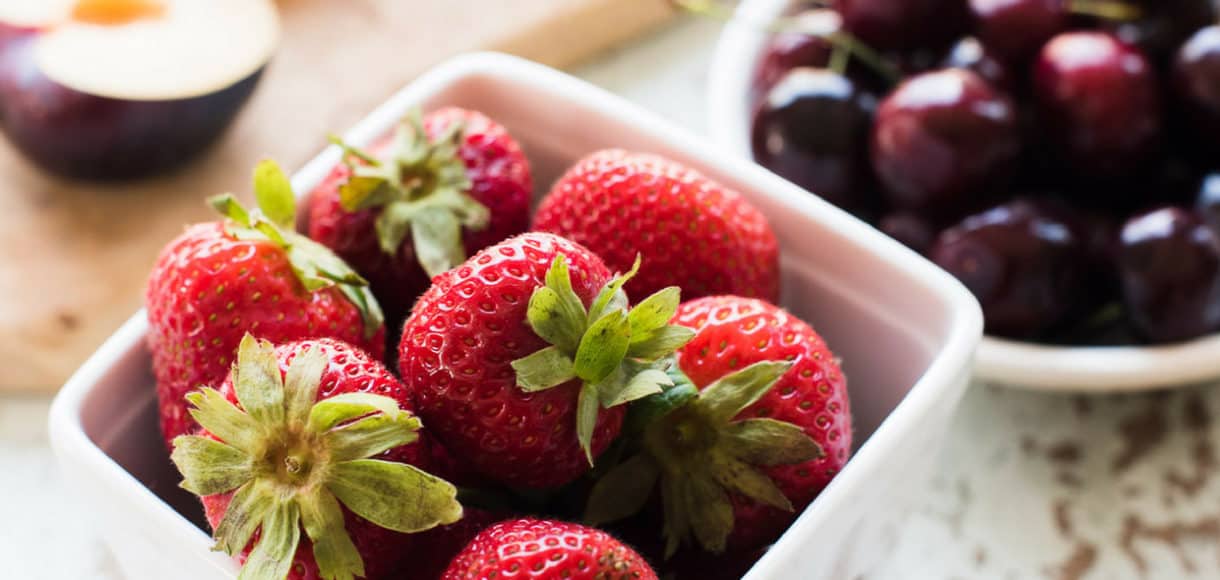 Strawberries in a white ceramic basket, with cherries and plums in the background.