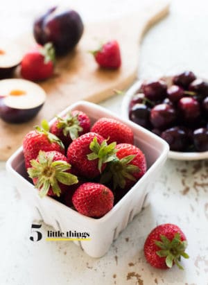 Strawberries in a white ceramic basket, with cherries and plums in the background.