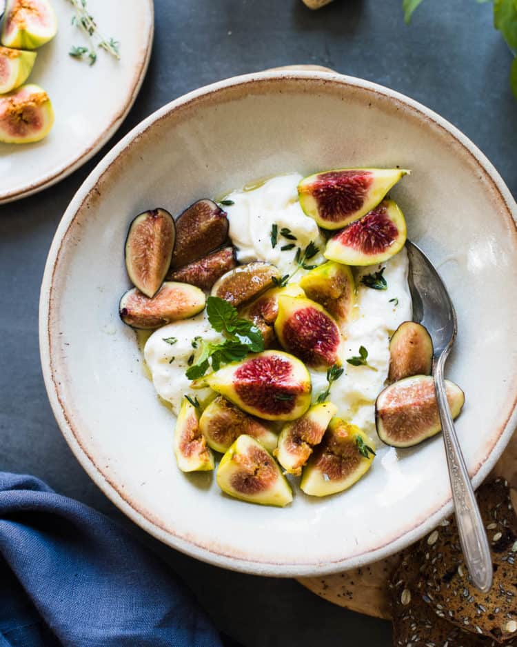 Cream bowl with torn burrata, sliced figs, herbs and a honey-balsamic glaze in a recipe for Honey Balsamic Figs with Burrata Cheese.