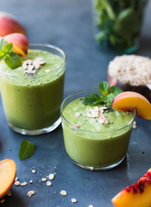 Peaches and Cream Green Breakfast Smoothie in glasses with a wedge of fresh peach, sprig of mint and a sprinkling of oats and chia.