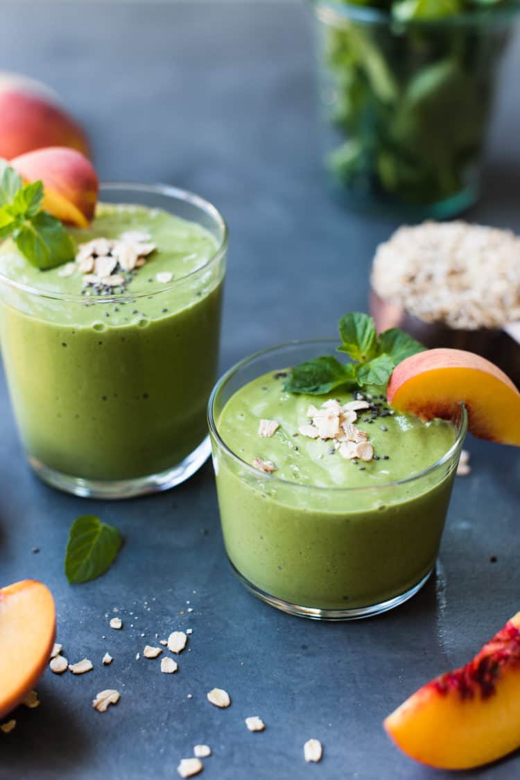 Peaches and Cream Green Breakfast Smoothie in glasses with a wedge of fresh peach, sprig of mint and a sprinkling of oats and chia.