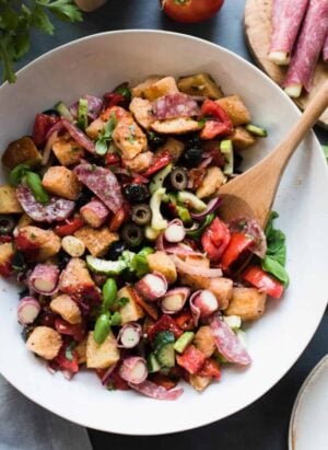 Muffuletta Panzanella Salad in a white bowl with a wooden spoon.