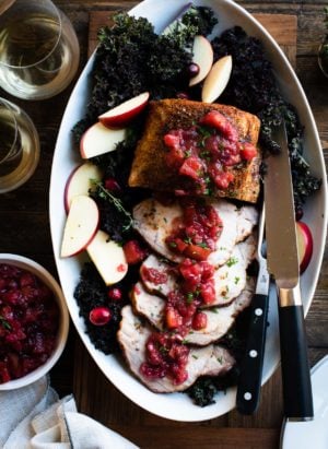 Roast Pork Loin with Apple Chutney sliced on a platter with fresh apple slices on a bed of kale. Glasses wine and a bowl of chutney on the side.